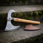 Vegvisir damascus axe with an open crescent bearded head, hooked horn and pale ash haft leaning on mossy stone steps beside its brown leather blade mask