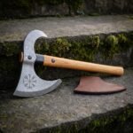 Vegvisir damascus axe with an open crescent bearded head, hooked horn and pale ash haft leaning on mossy stone steps beside its brown leather blade mask