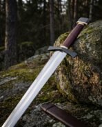 Viking arming sword with blackened cross guard and oxblood grip leaning on a mossy boulder beside its leather scabbard