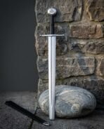 Viking arming sword with a rune column cut down the satin blade and a Valknut engraved disc pommel, standing against a stone wall beside a black leather scabbard