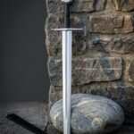 Viking arming sword with a rune column cut down the satin blade and a Valknut engraved disc pommel, standing against a stone wall beside a black leather scabbard