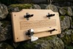 Viking axe wall plaque in carved oak with a Celtic knotwork band and leather lined steel cradles holding a hand forged bearded axe, propped on a mossy stone wall