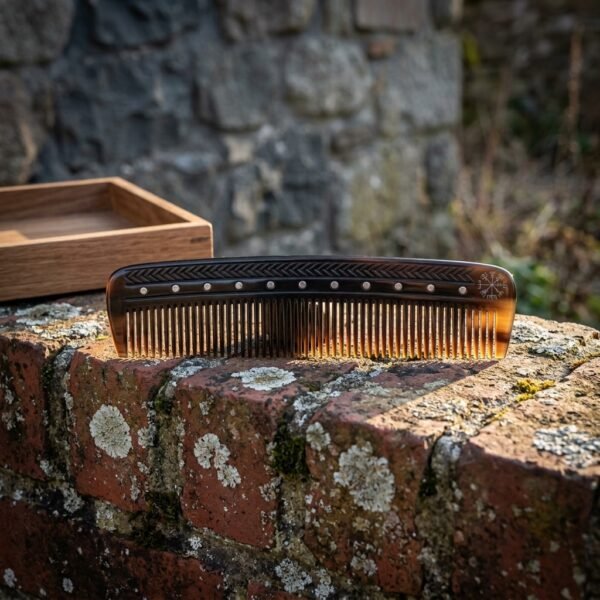 Viking beard comb cut from amber and dark natural ox horn with a Vegvisir stave and dotted spine, resting on a lichened brick wall beside its wooden box