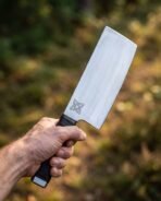 Viking cleaver with tall straight blade, engraved Celtic knot and dark wrapped handle held up in one hand outdoors