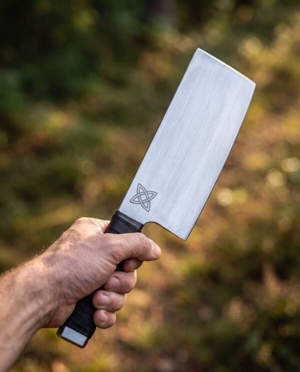 Viking cleaver with tall straight blade, engraved Celtic knot and dark wrapped handle held up in one hand outdoors