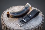 Viking drinking horn in polished amber and grey ox horn with steel rim band, shown on a weathered tree stump beside a black leather belt hanger with engraved raven