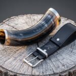 Viking drinking horn in polished amber and grey ox horn with steel rim band, shown on a weathered tree stump beside a black leather belt hanger with engraved raven