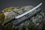 Viking katana with a Valknut etched 1095 steel blade and black leather wrapped tsuka resting on a lichen covered boulder beside its charcoal lacquered saya