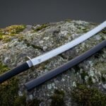 Viking katana with a Valknut etched 1095 steel blade and black leather wrapped tsuka resting on a lichen covered boulder beside its charcoal lacquered saya