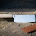 Viking kitchen cleaver with a flat stainless nakiri style blade, cream cord wrapped handle and engraved knotwork panel resting on a worn stone step