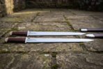Viking langseax pair with leather grips laid flat on weathered flagstones, full length view with scabbards to the right