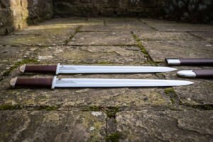 Viking langseax pair with leather grips laid flat on weathered flagstones, full length view with scabbards to the right
