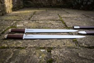 Viking seax set of two long single edged blades in polished 1095 steel with dark brown leather grips and steel caps, laid on worn flagstones beside their scabbards