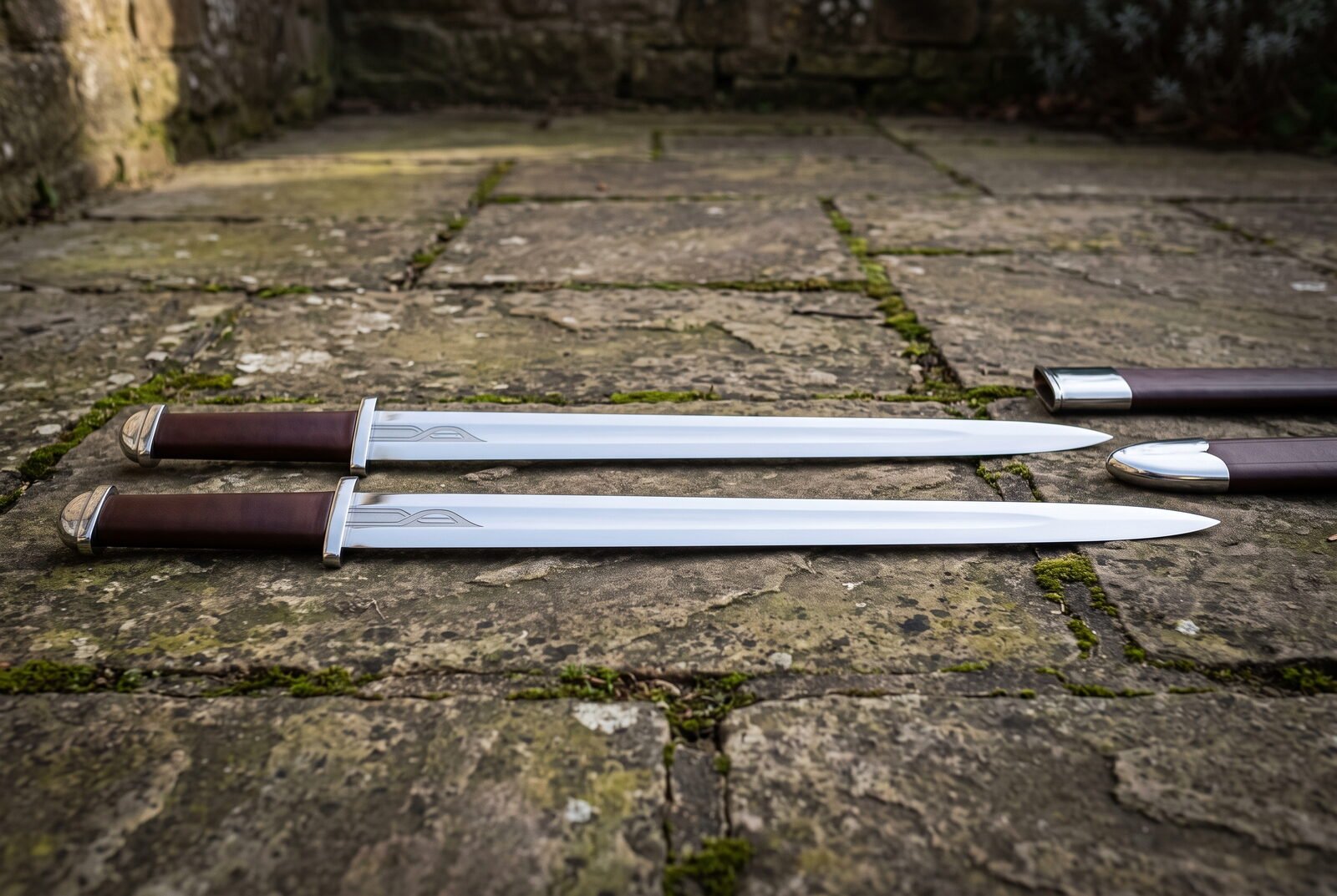 Viking seax set of two long single edged blades in polished 1095 steel with dark brown leather grips and steel caps, lai Viking seax set of two long single edged blades in polished 1095 steel with dark brown leather grips and steel caps, laid on worn flagstones beside their scabbards