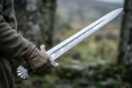 Viking short sword with etched interlace on the blade and lobed pommel held in a gloved hand outdoors