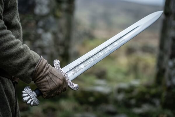 Viking short sword with etched interlace on the blade and lobed pommel held in a gloved hand outdoors