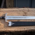 Viking short sword with a five lobe pommel and etched knotwork blade resting on split oak beside a black leather scabbard mounted with a bright triquetra throat