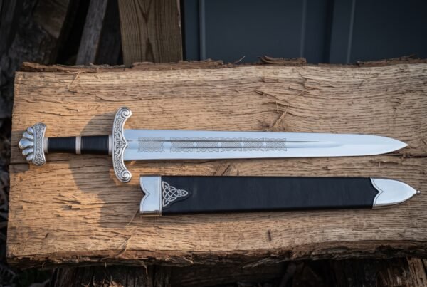 Viking short sword with a five lobe pommel and etched knotwork blade resting on split oak beside a black leather scabbard mounted with a bright triquetra throat