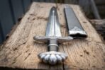 Viking short sword with five lobe pommel, knotwork guard and black grip beside its black leather scabbard on a plank