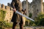 Viking short sword with triple fullered blade and downswept guard held by a man in front of castle ruins
