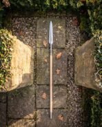 Viking spear with long leaf shaped steel head and steel butt ferrule laid on a flagstone path between mossy stone pillars