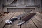 Viking sword with a five lobed steel pommel and black leather grip laid on weathered planks by an old plank door, brown leather scabbard alongside