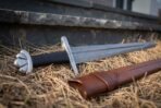 Viking sword with rune etched steel blade and five lobed pommel resting on dry straw against a stone wall, brown leather scabbard beside it
