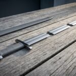 Viking sword with an elder futhark rune line cut above the guard, grey leather grip and rounded pommel resting on weathered decking beside its dark scabbard