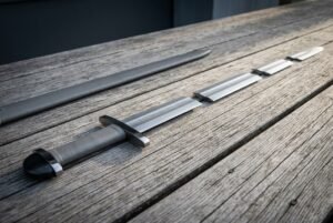 Viking sword with an elder futhark rune line cut above the guard, grey leather grip and rounded pommel resting on weathered decking beside its dark scabbard