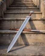 Viking sword with chevron pattern fuller, brown leather grip and steel guard propped on worn steps beside its scabbard