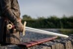 Viking sword with fullered steel blade and brass hilt held point down by a man in a leather coat, red scabbard on the wall