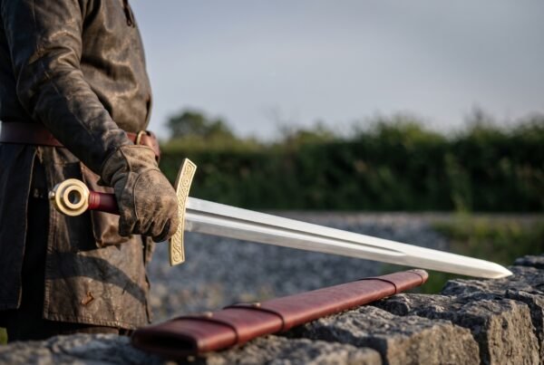 Viking sword with fullered steel blade and brass hilt held point down by a man in a leather coat, red scabbard on the wall