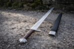 Viking sword with lobed steel pommel, brown leather grip and etched knotwork panel lying on gravel next to a black scabbard