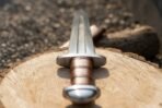 Viking sword with rune inscribed fuller, russet leather grip and domed steel pommel lying on a sawn log, low end view