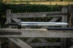 Viking sword with Valknut engraved black guard and leather wrapped grip resting on a wooden field gate with its black scabbard