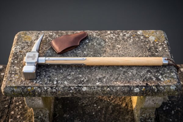 Viking war hammer with a forged square face, curved beak and Mjolnir engraved head on a pale ash haft with steel langets, lying on stone beside its leather head cover