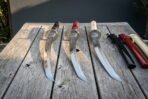 Wakizashi sword set of three hand forged 1095 steel blades with black red and ivory cord handles resting on a weathered wooden table