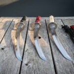 Wakizashi sword set of three hand forged 1095 steel blades with black red and ivory cord handles resting on a weathered wooden table