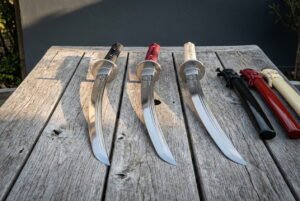 Wakizashi sword set of three hand forged 1095 steel blades with black red and ivory cord handles resting on a weathered wooden table