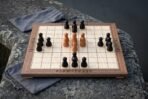 Walnut and maple hnefatafl board game with turned wood playing pieces, an Elder Futhark rune border and a cotton drawstring pouch, set on a lichen covered stone