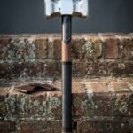 Walnut haft war hammer with a double faced polished steel head, engraved blackened collar and black leather grip standing upright against a mossy red brick wall