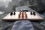 Walnut and maple hnefatafl board game with pieces mid game on a rocky ledge in fog, a grey drawstring bag in front