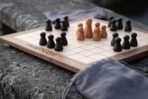 Walnut and maple hnefatafl board with turned dark and pale wooden pieces and a rune border, set on a stone wall with a grey cloth bag