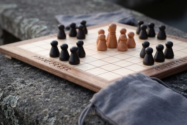 Walnut and maple hnefatafl board with turned dark and pale wooden pieces and a rune border, set on a stone wall with a grey cloth bag
