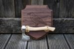 Walnut shield plaque engraved with a raven holding a polished bearded axe on brass brackets, leaning on a timber fence