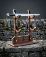 Walnut sword display stand with brass lined cradles and a cut Mjolnir engraving holding a Viking sword, photographed on a stone ledge before a dry stone wall