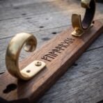 Close up of a walnut sword wall mount with polished brass scroll hooks and a row of runes engraved along the timber
