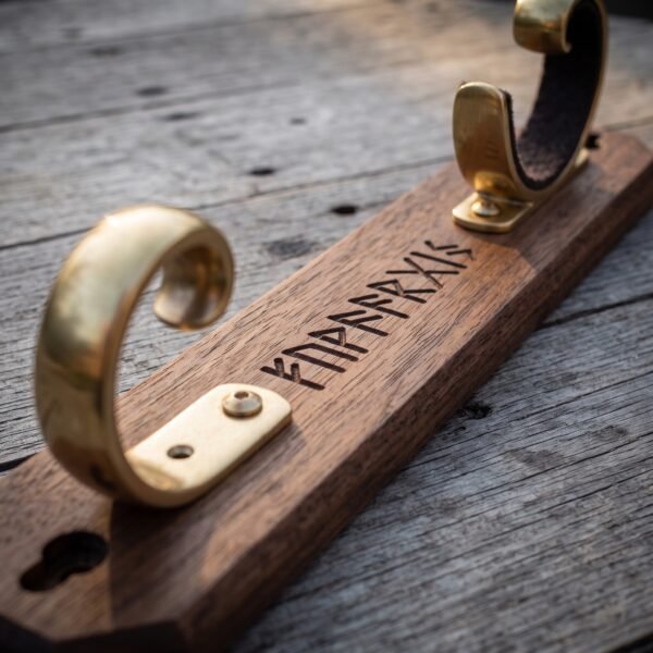 Close up of a walnut sword wall mount with polished brass scroll hooks and a row of runes engraved along the timber