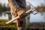 War hammer with Mjolnir engraved steel head and ash haft gripped in a leather gloved hand beside a lake