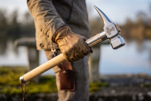 War hammer with Mjolnir engraved steel head and ash haft gripped in a leather gloved hand beside a lake